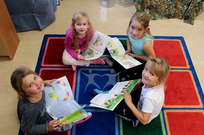 four students sitting on a carpet reading books and looking at the camera smiling.
